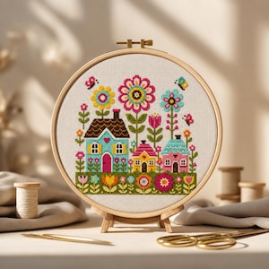 May include: A colorful cross-stitch embroidery of a whimsical village scene. The design features several small houses with vibrant colors, surrounded by large flowers and butterflies. The embroidery is displayed in a wooden hoop, perfect for home decor.