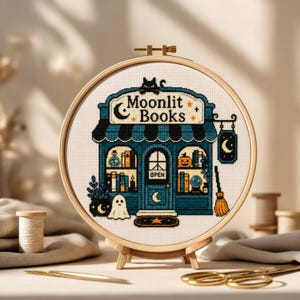 May include: A circular wooden embroidery hoop containing a cross-stitch design of a bookshop storefront titled Moonlit Books. The shop features a black cat, a ghost, a broom, a pumpkin, and books in blue and orange tones on a white fabric background.