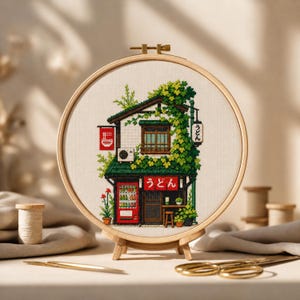May include: A cross-stitch embroidery of a Japanese udon shop in a wooden hoop. The shop features a red vending machine, green roof, and the Japanese characters for udon. The embroidery is displayed on a wooden stand.