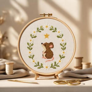 Puede incluir: Un bastidor de bordado circular de madera que contiene un patrón de punto de cruz sobre tela blanca. El diseño presenta un ratón marrón centrado dentro de una corona ovalada de margaritas blancas con hojas verdes y una estrella amarilla.