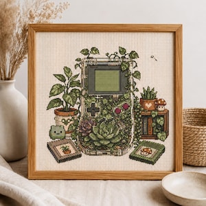 May include: A cross-stitch embroidery in a light wood frame featuring a handheld gaming console overgrown with green vines and succulents. The design includes potted plants, a small frog figure, and two game cartridges with plant and mushroom icons.