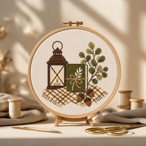 Puede incluir: Un bastidor de bordado circular de madera que contiene un patrón de punto de cruz sobre tela blanca. El diseño presenta una linterna marrón, un libro verde con un motivo de hoja blanca, una rama frondosa, dos bellotas y una manta a cuadros.