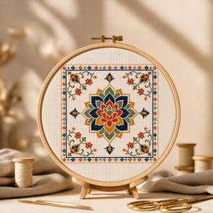 Puede incluir: Un bastidor de bordado circular de madera que contiene una tela blanca con un diseño de mandala en punto de cruz. El patrón presenta una flor central similar a un loto en tonos azul marino, naranja, amarillo y verde salvia, enmarcada por un borde.
