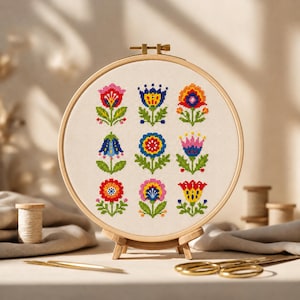 May include: A colorful cross-stitch floral design in a wooden embroidery hoop. The design features nine stylized flowers in various colors, including pink, blue, orange, and red, on a cream-colored fabric. The hoop is displayed on a small wooden stand.