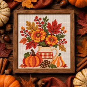 May include: A framed cross-stitch embroidery featuring a bouquet of orange and yellow flowers, red maple leaves, and berries in a cream-colored pitcher. Beside the pitcher are a round orange pumpkin, a small striped gourd, and a brown pinecone.