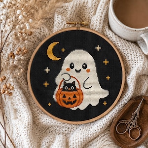 May include: A circular wooden embroidery hoop containing a cross-stitch design on black fabric. The design features a white ghost holding an orange jack-o'-lantern with a small black cat inside, surrounded by a yellow crescent moon and stars.