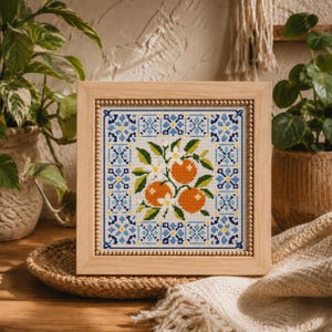 May include: A square cross-stitch embroidery in a light wood frame with a beaded inner border. The design features three orange fruits with green leaves and white blossoms, surrounded by a symmetrical blue and yellow geometric tile pattern.