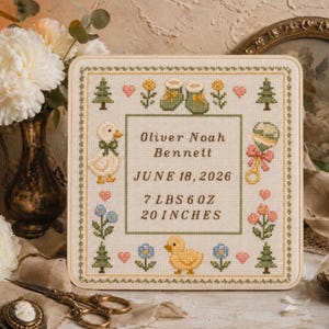 May include: A square cross-stitch sampler on cream fabric featuring a decorative border of pine trees, flowers, hearts, baby booties, a duck, and a rattle. The center text reads: Oliver Noah Bennett, JUNE 18, 2026, 7 LBS 6 OZ, 20 INCHES.