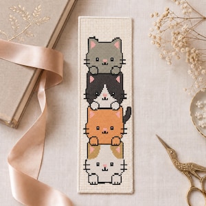 May include: A rectangular cross-stitch bookmark on cream-colored fabric featuring a vertical stack of four stylized cats. The cats are colored gray, black and white, orange, and white with brown patches, each with simple facial features and small pink ears.