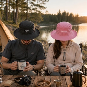 May include: A black bucket hat with the name Kyle embroidered in white and a pink bucket hat with the name Emma embroidered in white. Both hats feature a wide brim, a chin strap with an adjustable toggle, and a structured crown design.