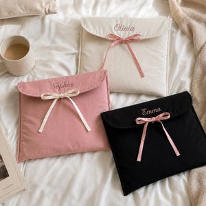 May include: Three rectangular fabric pouches with envelope-style flaps, each featuring a ribbon tie closure and embroidered names. One white pouch reads Olivia, one pink pouch reads Sophia, and one black pouch reads Emma in cursive script.