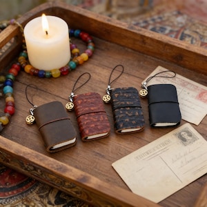 May include: Four miniature leather journals in shades of brown and black, each with a charm and a black cord. A lit white candle and a beaded necklace are in the background, all on a wooden tray. Two vintage postcards are also present.