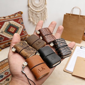 Op de afbeelding: Een verzameling miniatuur leren notitieboekjes in verschillende bruin- en zwarttinten, vastgehouden in een hand. Elk notitieboekje is vastgemaakt met een touwtje en heeft de initialen "A.K." gegraveerd. Sommige hebben decoratieve bedels.