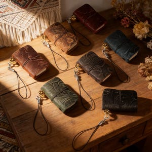 May include: A collection of small, leather-bound journals in shades of brown, green, and black. Each journal has an embossed floral pattern and a wrist strap with a gold-coloured charm. The journals are displayed on a wooden surface.