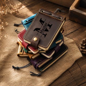 May include: A stack of small leather-bound notebooks in various colors including brown, blue, red, green, and purple. Each notebook features a metal snap closure, visible stitching, and a cord tie. The top brown notebook is engraved with the text Master.