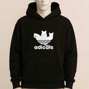 May include: Black hooded sweatshirt with a white graphic of three cats above the text "adicats". The sweatshirt has a front pocket and a drawstring hood. The design is a parody of a well-known athletic brand.