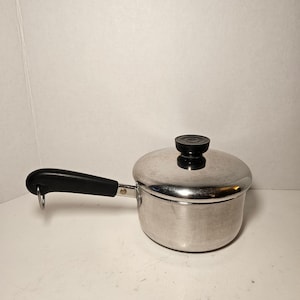 May include: A small, stainless steel saucepan with a lid and a black handle. The lid has a black knob. The handle has a loop for hanging. The pot is designed for cooking and is likely used in a kitchen.