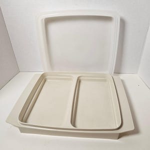 May include: A beige, square Tupperware container with a divided interior and a matching lid. The container has a rectangular shape with rounded corners and a raised edge. The lid is partially open, revealing the interior compartments.