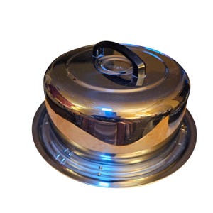 以下が含まれることがあります： ドーム型の蓋と黒いハンドルが付いた、光沢のあるステンレス製のケーキキャリア。キャリアは、お揃いの丸いベースの上にあります。蓋の反射面は周囲の環境を映し出しています。