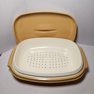 以下が含まれることがあります： 温かみのある黄色い色合いのヴィンテージTupperwareスチーマー。長方形で、穴あき白インサートが付いています。蓋は開いており、内部が見えます。スチーマーは調理と食品保存用に設計されています。