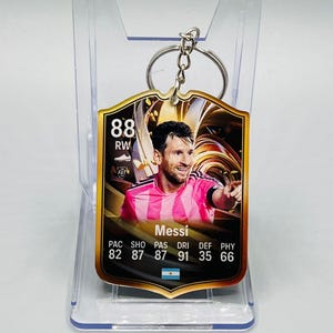 Op de afbeelding: Een sleutelhanger met een voetbalspeler-kaartontwerp. De kaart heeft een gouden rand en toont een foto van een voetballer in een roze en wit gestreept shirt. De kaart toont de naam van de speler, Messi, en diverse statistieken.