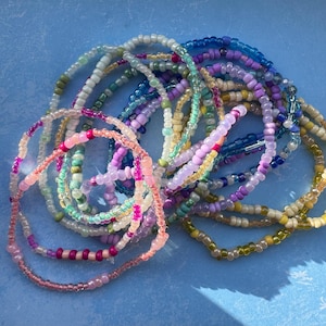 May include: A collection of multiple layered beaded bracelets featuring a variety of small, colourful glass seed beads in shades of pink, purple, blue, green, yellow, and clear. The bracelets are arranged in a tangled, circular cluster.
