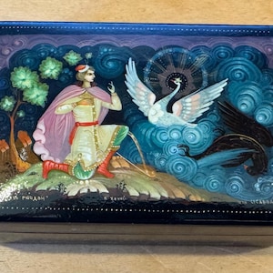 Puede incluir: Una caja lacada en negro con una pintura detallada. La obra de arte representa una figura con capa rosa, un cisne blanco y un cisne negro sobre un fondo de nubes azules arremolinadas y árboles verdes. Acentos dorados enmarcan la caja.