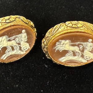 May include: Two oval-shaped gold-tone earrings. Each earring features a cameo with a brown background and a white relief carving of a chariot pulled by horses. The earrings have a decorative gold border.