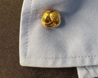 Puede incluir: Primer plano de un gemelo dorado en forma de nudo en una camisa azul claro. El gemelo tiene una superficie texturizada y está sujeto al puño de la camisa. La camisa está hecha de una tela finamente tejida.