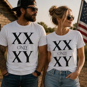 May include: Two people wearing white t-shirts with the text "XX ONLY XY" in black. The shirts are paired with blue jeans. The background includes a wooden structure and an American flag.