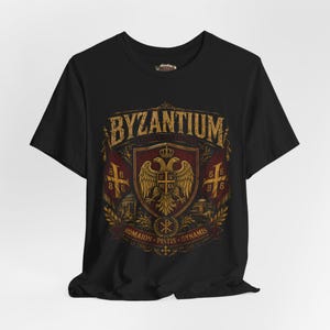 Può includere: T-shirt nera con grafica dorata invecchiata. Il design presenta la scritta "Byzantium", uno stemma con un'aquila bicipite, bandiere e testo in stile vintage.