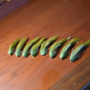 May include: Eight olive green fishing flies are arranged on a wooden surface. Each fly has a fluffy, textured body and a silver-colored accent. The flies are designed for fly fishing.