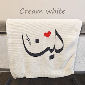 Peut inclure: Une couverture en tricot blanc crème avec une calligraphie arabe noire et une petite icône de cœur rouge. Le texte "Cream white" est imprimé au-dessus de la couverture. La couverture présente une surface tissée texturée avec un bord côtelé.