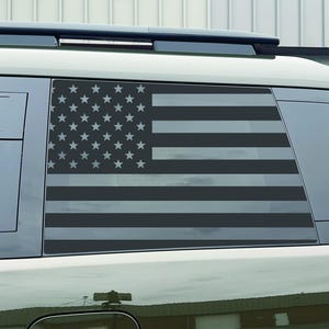 May include: A black and gray American flag design is displayed on the rear window of a vehicle. The flag features stars and stripes, creating a patriotic theme. The window decal is a decorative accessory.