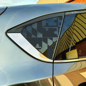 Puede incluir: Primer plano de una ventanilla de coche con un diseño de la bandera de Maryland. La bandera es un patrón geométrico de formas negras y grises. La ventana tiene un borde cromado y está colocada contra una carrocería gris metálico.