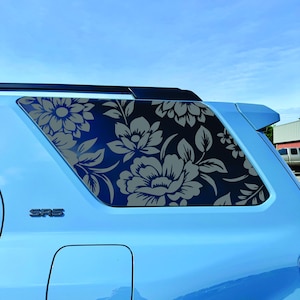 Peut inclure: Une voiture bleu clair avec un motif floral sur la vitre arrière. Le motif présente de grandes fleurs et des feuilles grises sur un fond sombre. La voiture porte les lettres "SRS" à l'arrière.