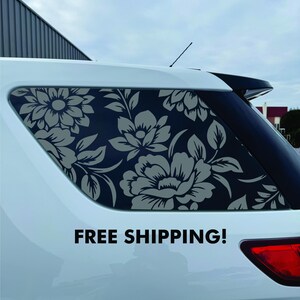 May include: A floral design car window decal. The decal features a pattern of large gray flowers and leaves against a dark background. The decal is applied to the rear window of a white car.