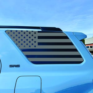 May include: A blue vehicle with a window decal featuring a stylized American flag design. The flag has gray stripes and blue stars on a gray background. The vehicle is parked outside on a sunny day.