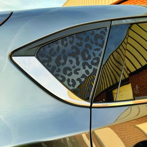May include: A close-up of a car's rear window featuring a dark gray leopard print design. The window is framed by a black border and a silver trim, reflecting the surrounding environment. The car's body is a metallic gray color.