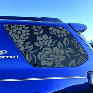 Peut inclure: Un autocollant floral gris foncé appliqué sur la vitre latérale arrière d'un véhicule bleu. L'autocollant présente un motif répétitif de grandes fleurs et de feuilles stylisées qui recouvrent la surface vitrée, contrastant avec la carrosserie.