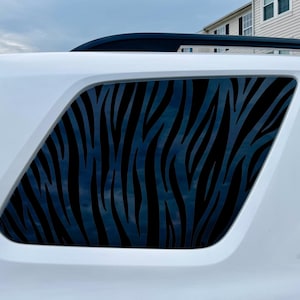 May include: White SUV with a rear window featuring a black zebra stripe pattern. The vehicle has a black roof rack and a dark-tinted rear window. The image is taken outdoors.