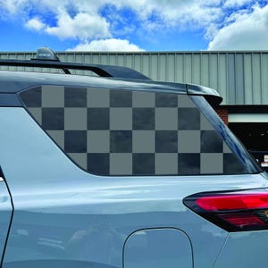 May include: A light grey vehicle rear quarter window featuring a dark grey and light grey chequered pattern decal. The decal covers the entire glass surface of the window, which is positioned below a black roof rack on a light grey car body.