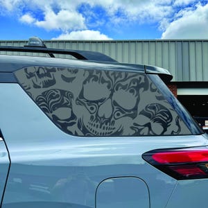 May include: A light grey vehicle rear side window featuring a dark grey decal with a repeating pattern of stylised skulls. The decal covers the entire glass surface, displaying intricate, flowing lines that form multiple skull shapes.
