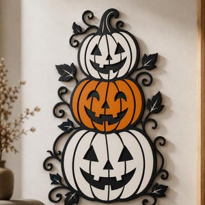 Puede incluir: Una decoración de pared con tres calabazas apiladas. Las calabazas superior e inferior son blancas, mientras que la del medio es naranja. Las tres tienen caras talladas en negro y están rodeadas de enredaderas y hojas decorativas negras.