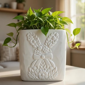 May include: A white ceramic planter with a raised bunny design filled with green leafy plants. The planter has a rectangular shape and a detailed embossed bunny with floral accents. The plant has heart-shaped leaves.