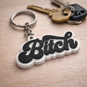 May include: A black and white keychain with the word "Bitch" in a bold, stylized font. The keychain is attached to a silver ring and sits on a wooden surface. Keys are visible in the background.