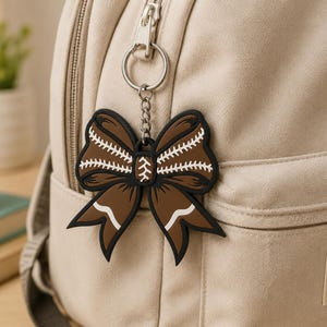 Peut inclure: Un porte-clés en caoutchouc marron et noir en forme de nœud avec des détails de coutures blanches style baseball. Le porte-clés est fixé à un anneau et une chaîne en métal argenté, accrochés à la fermeture éclair d'un sac à dos en toile beige.