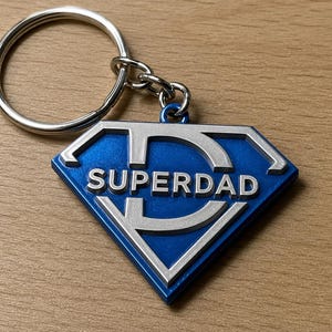 Peut inclure: Un porte-clés Superpapa bleu et argenté. Le porte-clés présente un pendentif en forme de diamant avec les mots "SUPERDAD" en lettres argentées. Le pendentif est attaché à un porte-clés argenté.