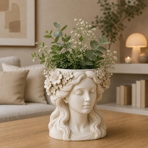 Peut inclure: Un pot décoratif crème en forme de tête de femme aux yeux fermés et aux cheveux ondulés. Le pot est rempli de feuillage vert et de fleurs blanches. La tête est ornée d'une couronne florale. Le pot est posé sur une table en bois.