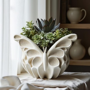 May include: A cream-colored butterfly-shaped planter filled with green succulents. The planter has detailed wing patterns and a rounded body. The background includes a window with a sheer curtain and a shelf with ceramic vases.